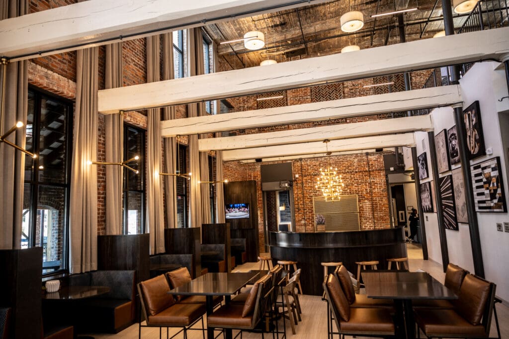 Modern restaurant interior with exposed brick walls, wooden beams, leather seating, and a central bar area with a hanging chandelier.