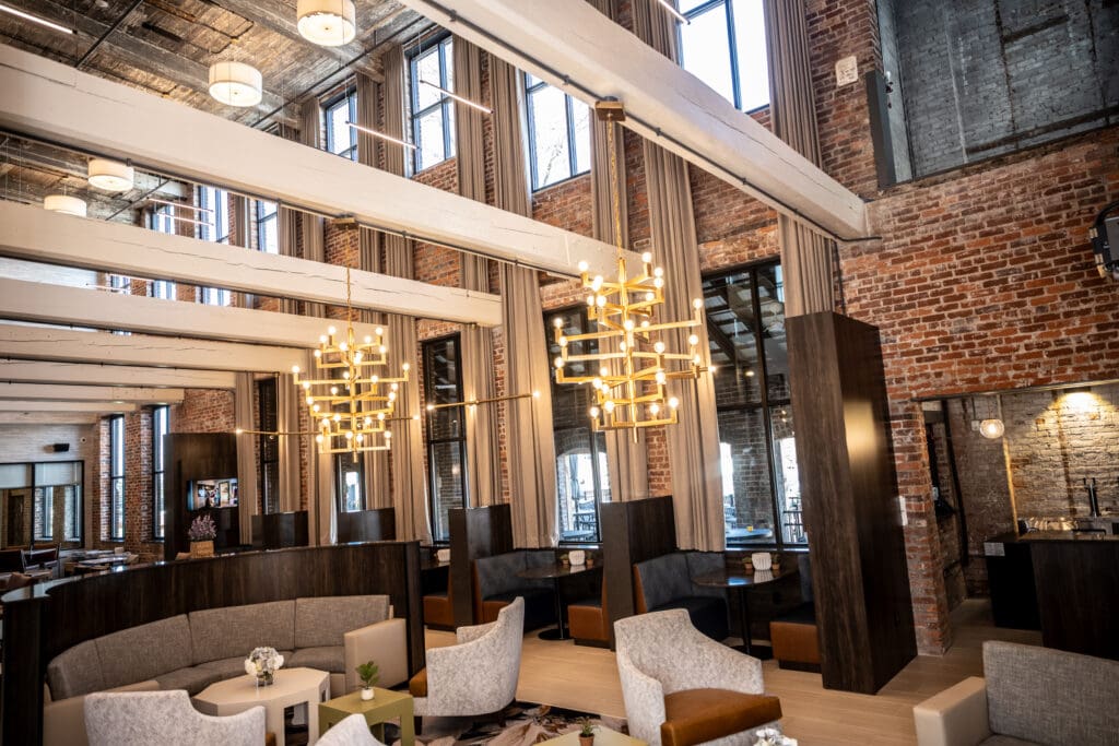 Modern lounge area with exposed brick walls, large windows, contemporary chandeliers, and assorted seating including armchairs and booth-style seats.