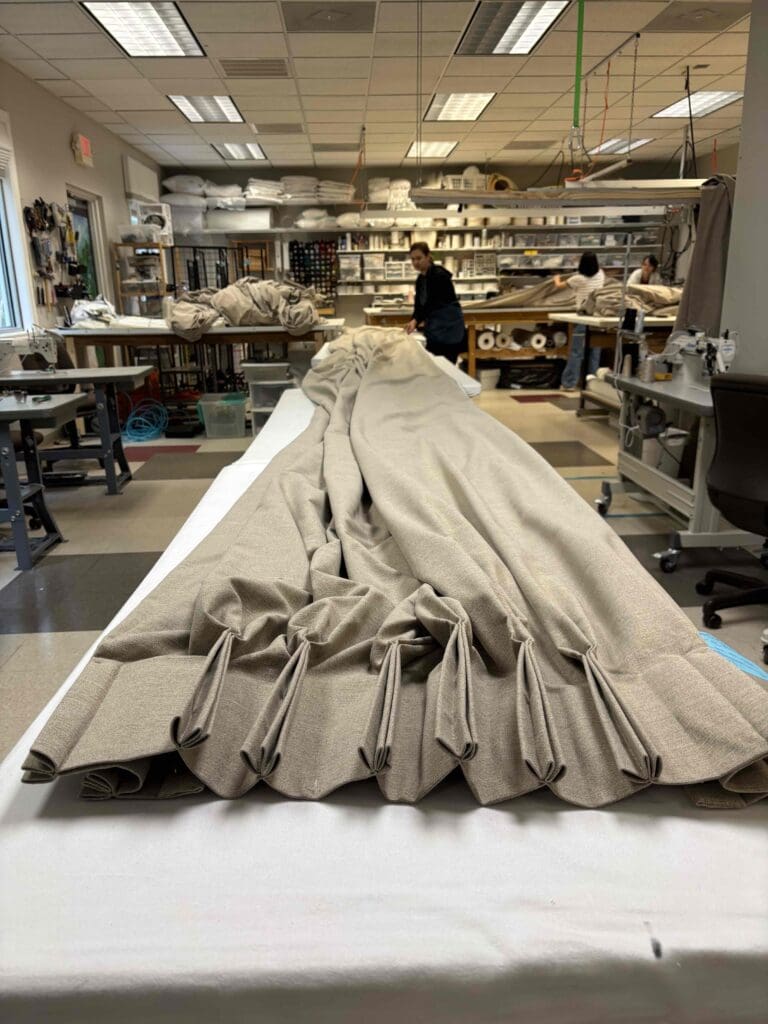 A long, beige curtain is laid out on a worktable in a fabric workshop, with workers and sewing equipment visible in the background.