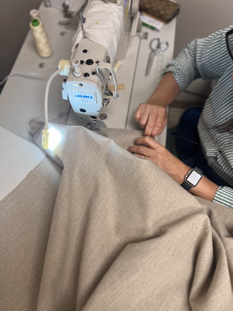A person using a sewing machine to stitch beige fabric, with sewing tools and threads nearby on the table.