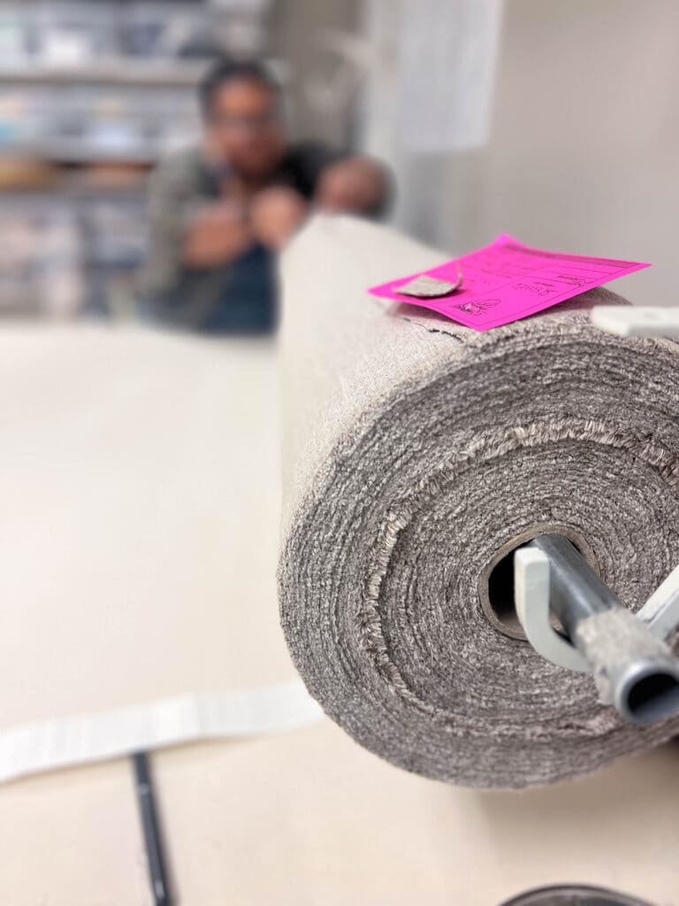 A large roll of gray fabric sits on a table with a pink tag attached; a person is blurred in the background, working.