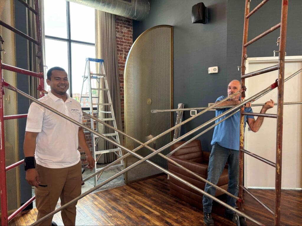 Two men stand among metal scaffolding poles in an indoor space with brick walls, wood floors, a ladder, and a large window in the background.