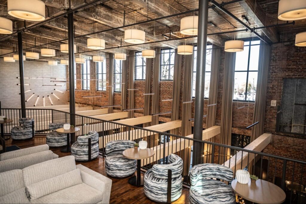 A modern loft lounge with exposed brick walls, large windows, wood beams, patterned chairs, white sofas, and hanging ceiling lights.