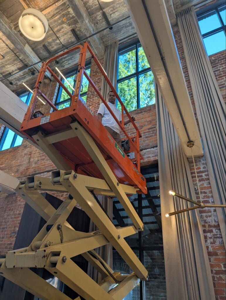 A person stands on an elevated scissor lift near tall windows inside a brick-walled room with high ceilings and exposed beams.