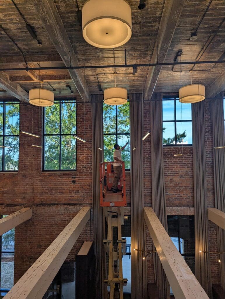 A worker on a lift performs maintenance on ceiling light fixtures in a tall, industrial-style room with brick walls and large windows.
