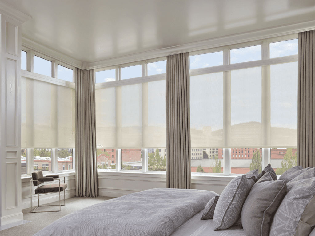 A modern bedroom with large windows covered by light-filtering roller shades, neutral-colored curtains, a neatly made bed, and a chair by the window.