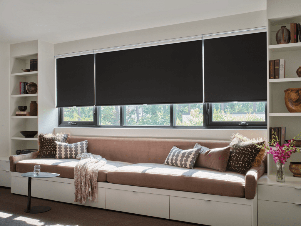 A modern living room with a long beige sofa, patterned cushions, built-in shelves, a round side table, and three large windows covered by black roller shades.
