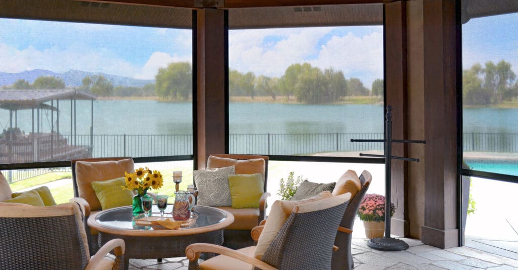 A covered patio with cushioned chairs, a round glass table, and decorative plants overlooks a lake and trees through mesh screens.