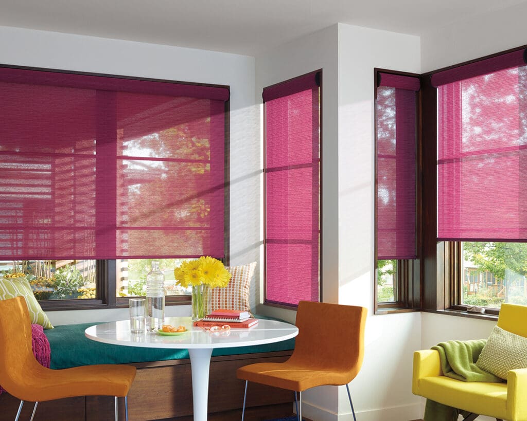 A modern dining area with a round white table, two orange chairs, a green armchair, and windows covered by magenta roller blinds. A vase of yellow flowers is on the table.