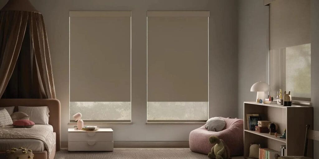 A softly lit bedroom with two large windows with beige roller blinds, a bed, a white nightstand, a pink bean bag, a turtle plush, and a bookshelf.