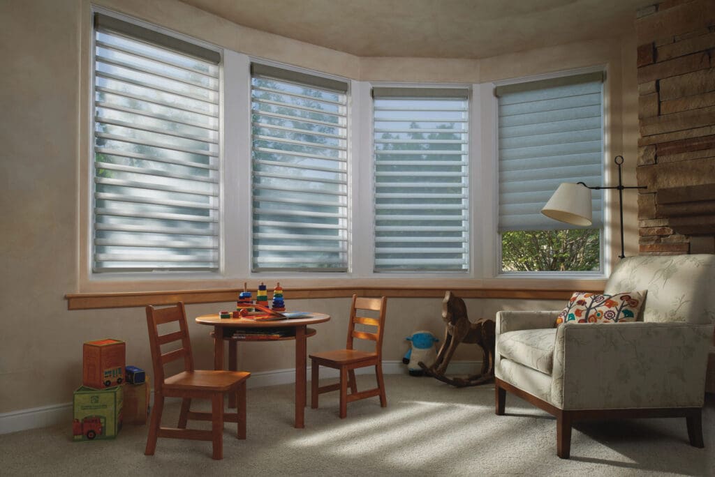 A sunlit playroom with a round table, three wooden chairs, toys, a rocking horse, an armchair, and large windows with blinds.