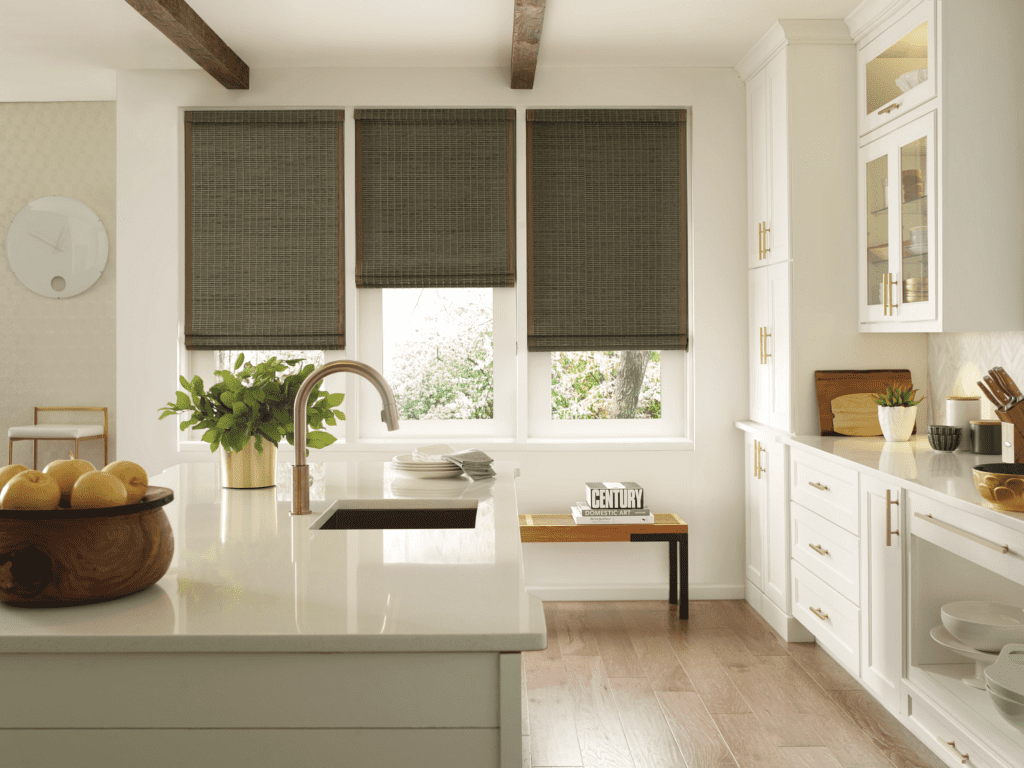 Modern kitchen with white cabinets, a center island with a sink, wooden bowl of lemons, potted plant, and three large windows covered with woven dark blinds.