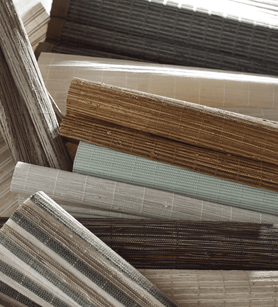 Rolled-up bamboo and woven blinds in various neutral and earth tones, including beige, brown, cream, and light blue, are stacked in a pile.