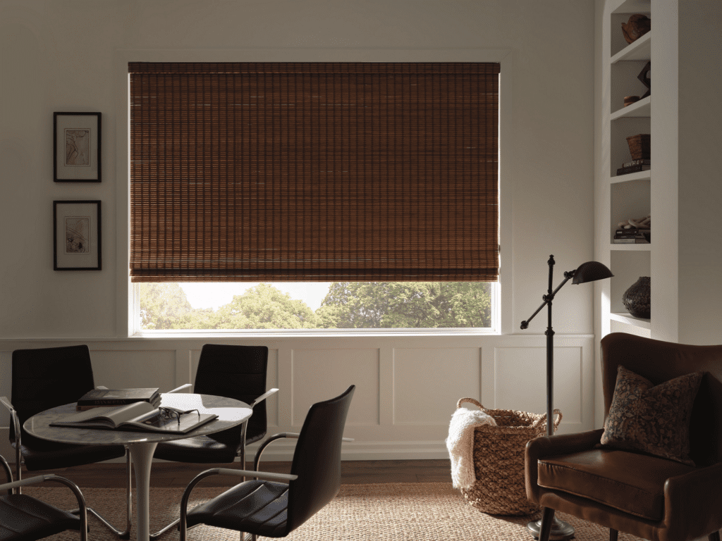 A room with a round table, four chairs, an armchair, a floor lamp, bookshelves, and a large window covered by a brown bamboo blind, letting in filtered light.
