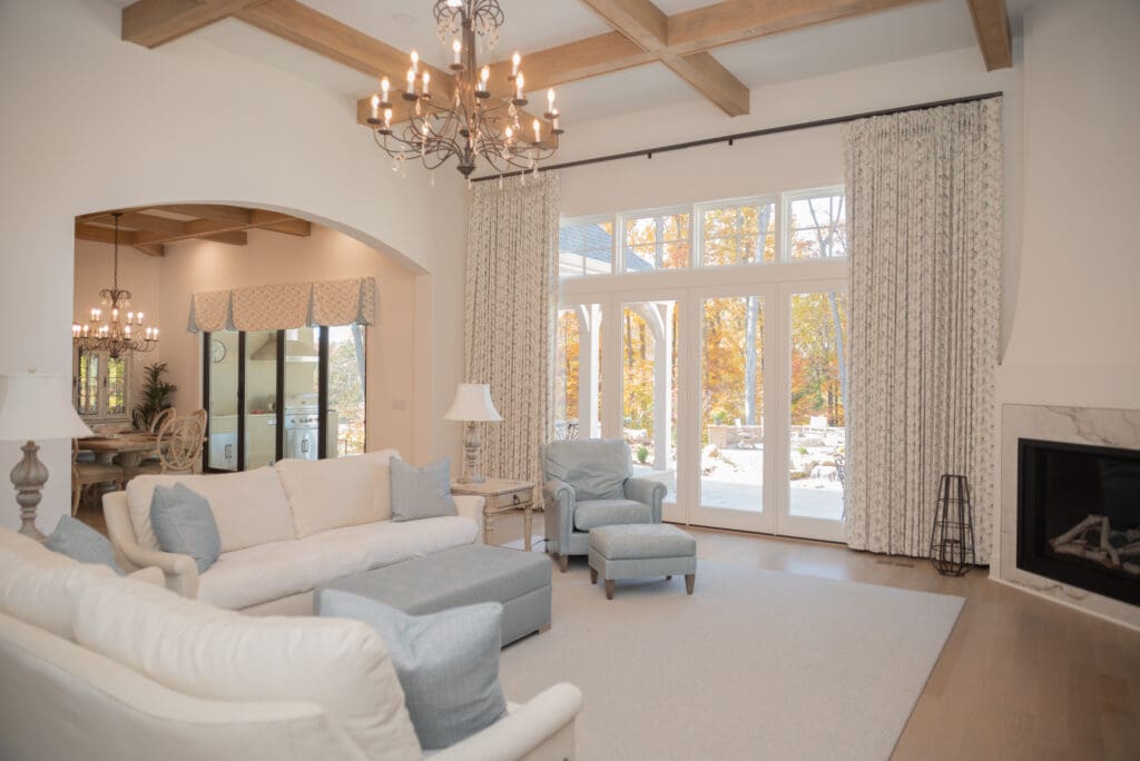 Bright living room with white sofas, blue armchairs, chandelier, fireplace, and large windows with curtains overlooking a patio and trees with autumn foliage.