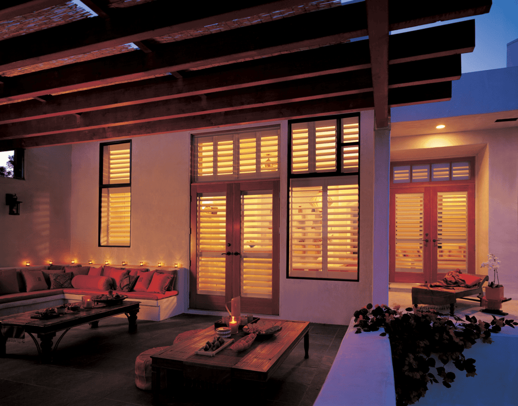 Outdoor patio with wooden furniture and cushions, under a pergola, facing a house with large windows and doors illuminated from inside. Candles are placed along the ledges and tables.