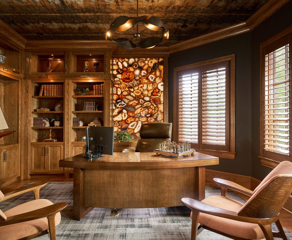 A home office with wood paneling, a large desk, bookshelves, two chairs, a computer monitor, window blinds, and a decorative stone art piece on the wall behind the desk.