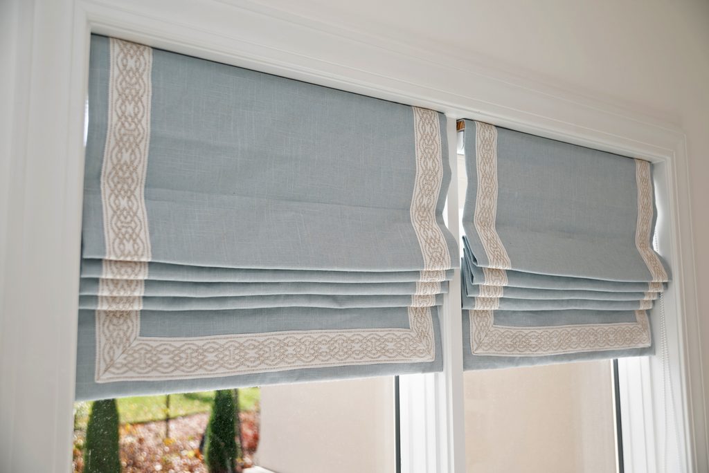 Two windows with light blue Roman shades featuring a decorative white border, partially raised to let in natural light.