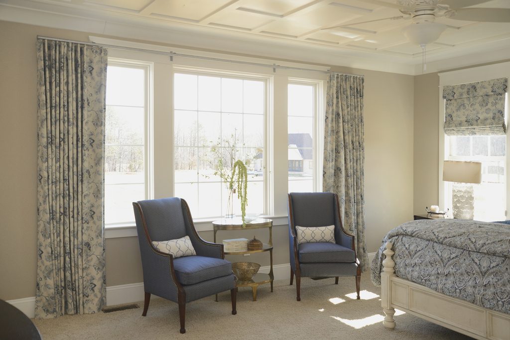 Bedroom with two blue armchairs, a small round table, patterned curtains, and large windows letting in natural light. Bed and lamp are partially visible on the right.