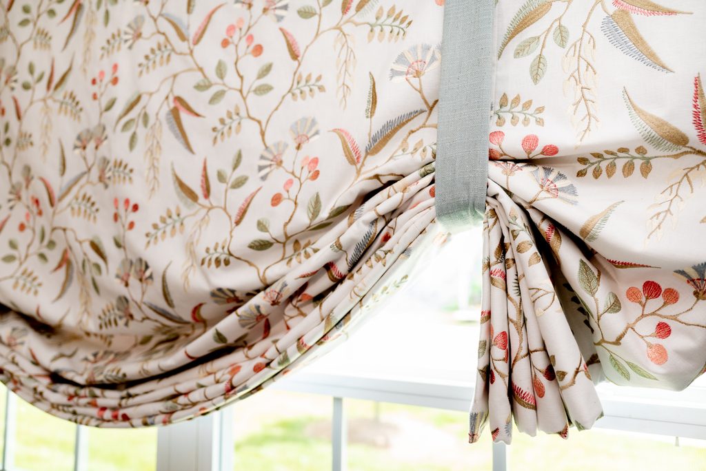 Close-up of a Roman shade with a botanical pattern in muted colors, partially raised and tied with a fabric strap, hanging over a window.
