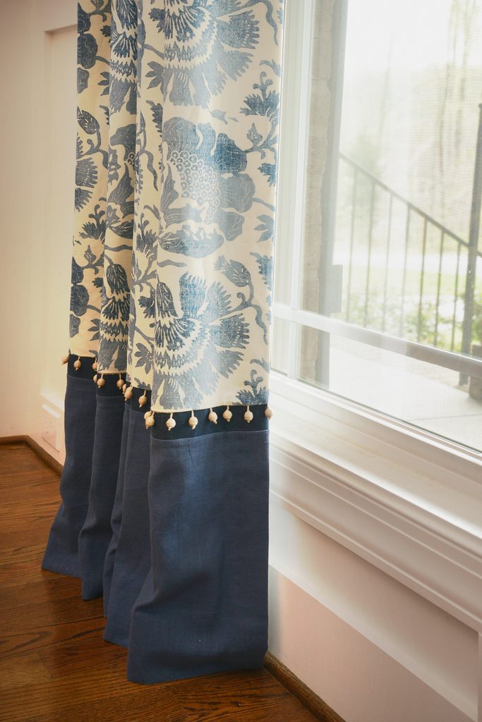 Blue and white floral curtains with tassels hang beside a window, partially open to reveal a view of steps and greenery outside.