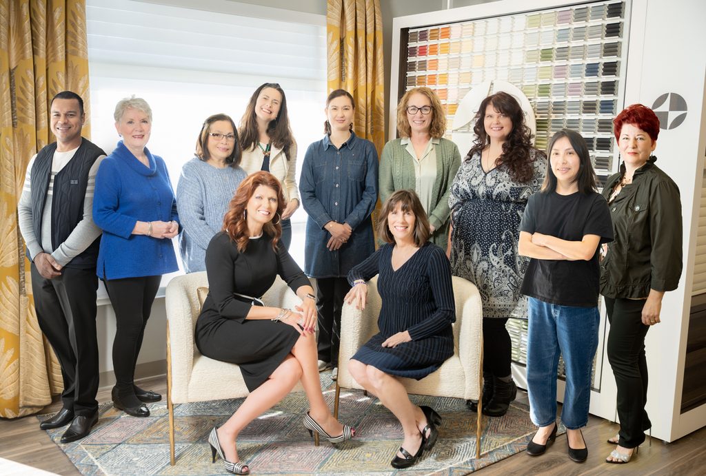 Twelve people pose indoors by a window and color sample wall, with two women seated in front and ten people standing behind them, all smiling for the camera.