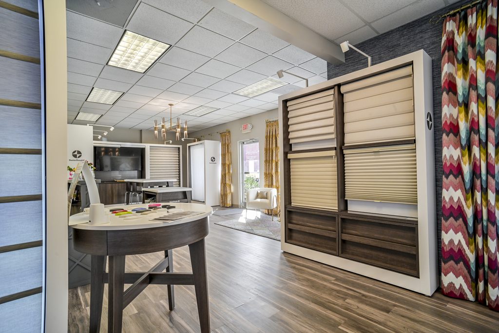 Showroom interior featuring window blinds displays, a table with fabric samples, wood flooring, and colorful patterned curtains. Large windows and ceiling lights provide ample illumination.