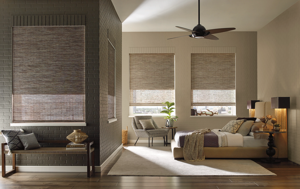 A bedroom featuring a bed, an armchair, and a bench, all accented by light-filtering woven wood shades on three windows.
