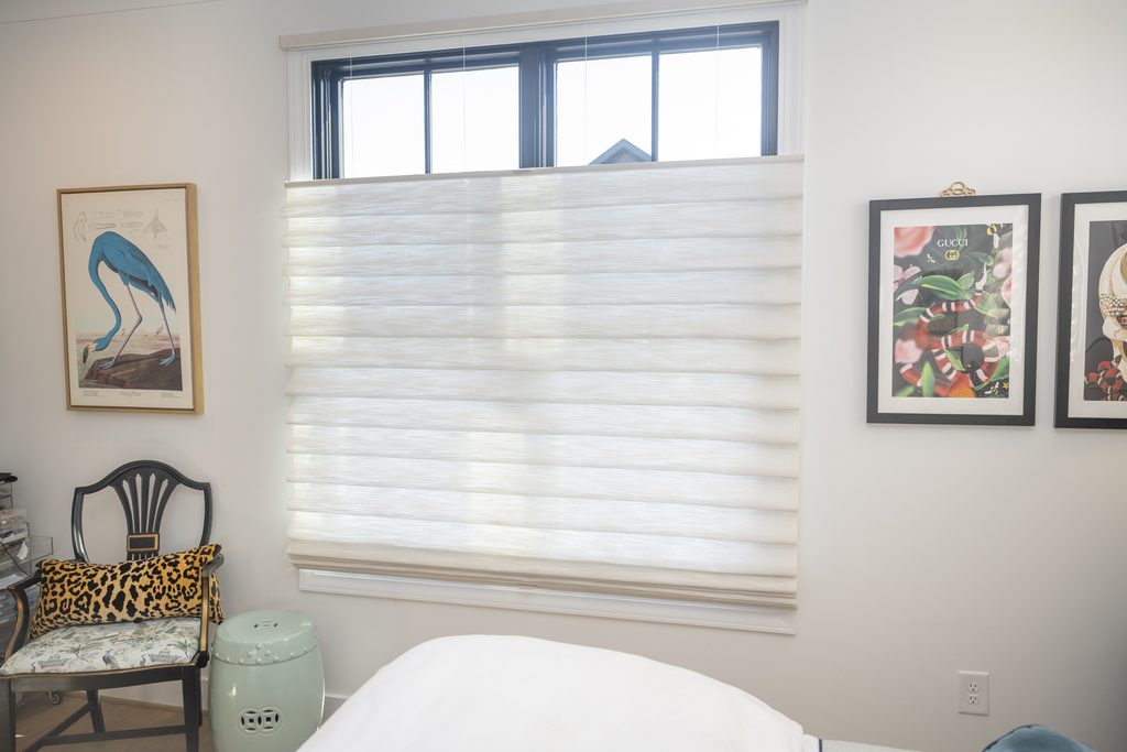 A white window with a pleated roman shade, flanked by two framed art prints and a chair with a leopard-print pillow.